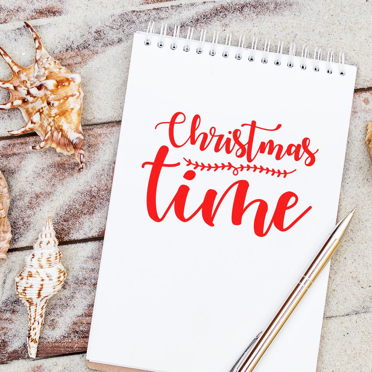 Red Christmas time stamp design on white spiral notebook, with seashells and gold pen on sandy wood background.