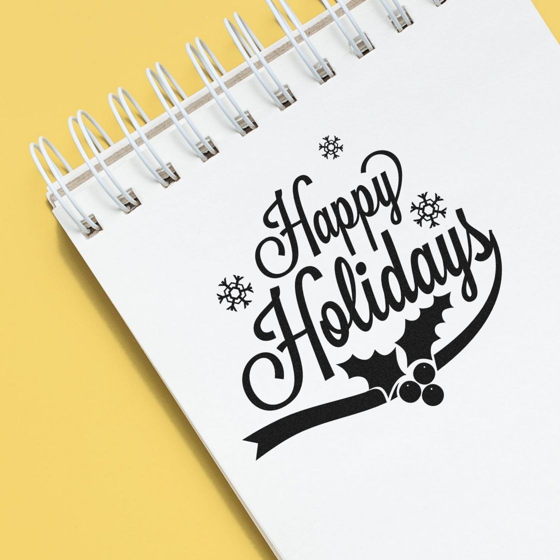 Black Happy Holidays stamp and seal design on white spiral notebook with snowflakes, holly, and yellow background.