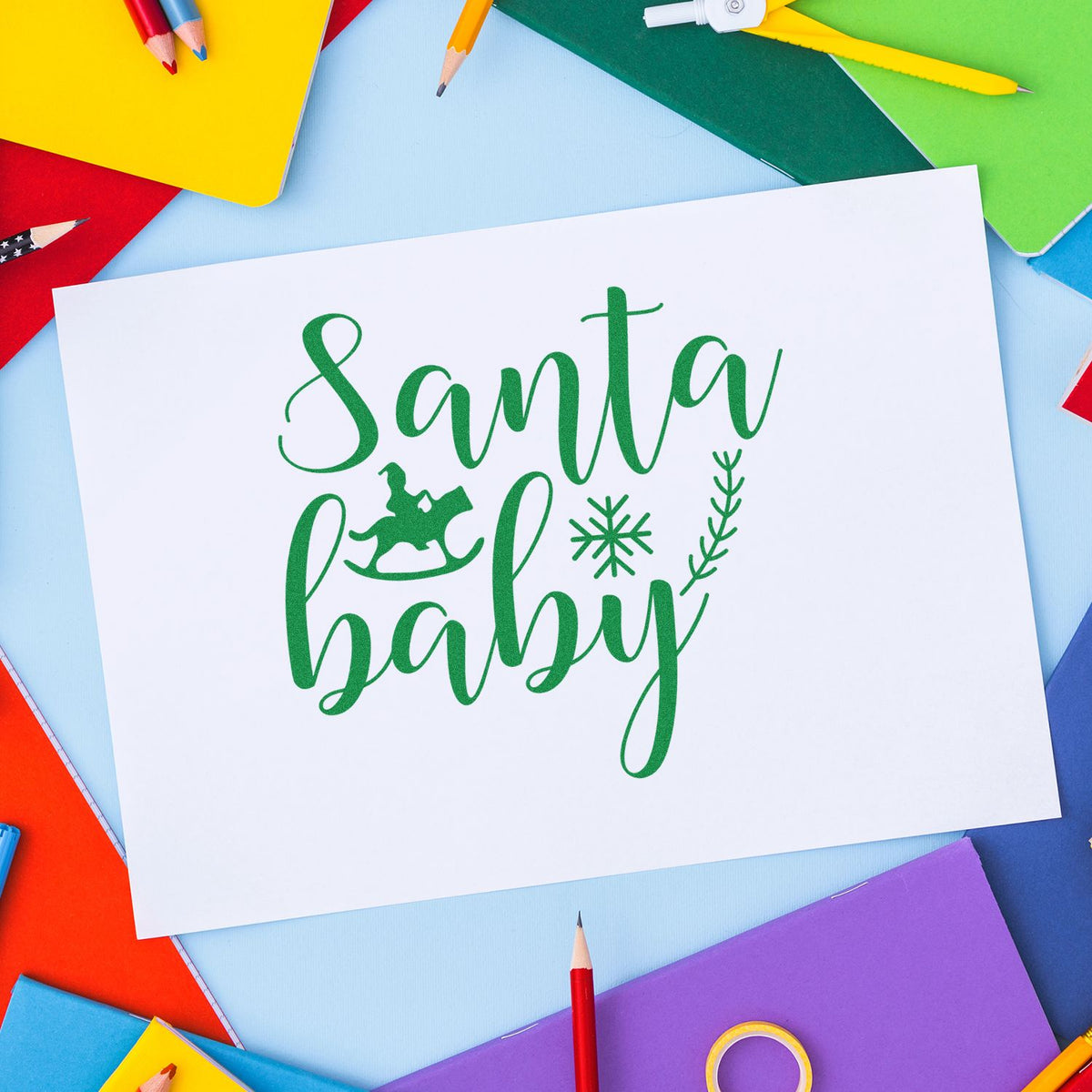 Green Santa Baby holiday stamp and seal with rocking horse, snowflake and wreath on white paper, surrounded by colorful notebooks and pencils.