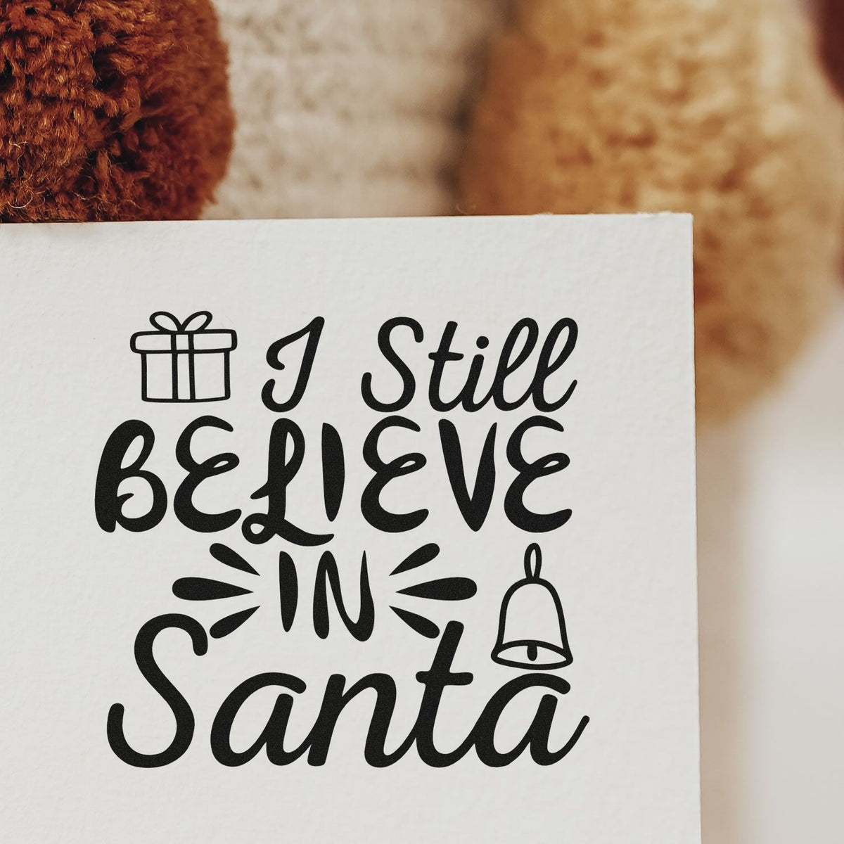 Black holiday stamp and seal on white paper reading I Still Believe In Santa, with gift and bell icons.