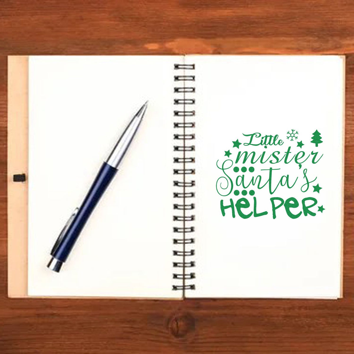 Open notebook with blue pen and green holiday stamp/seal reading Little mister Santas helper, with stars and tree.