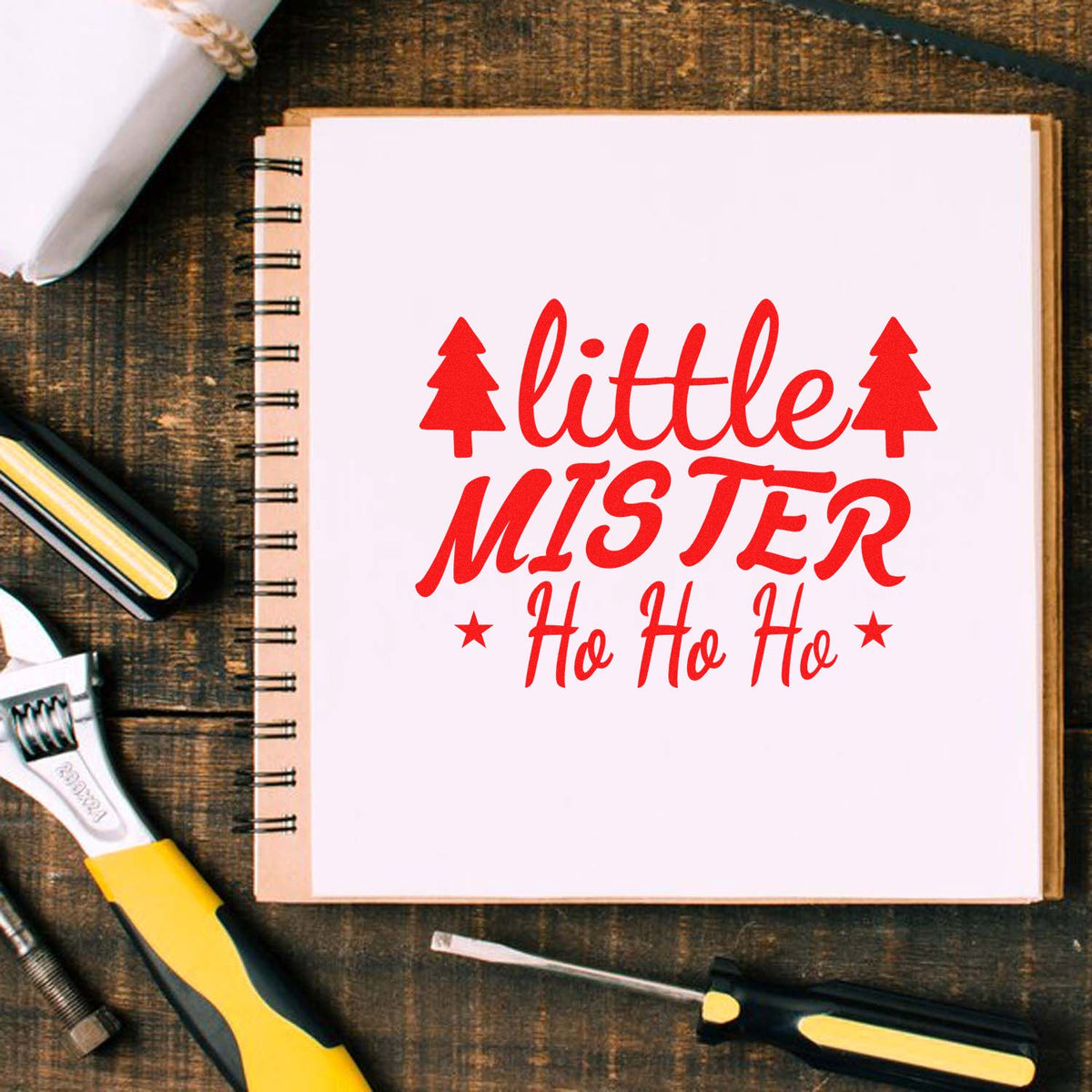 Red Little Mister Ho Ho Ho holiday stamp and seal on white spiral notebook, surrounded by yellow-black craft tools on rustic wood.