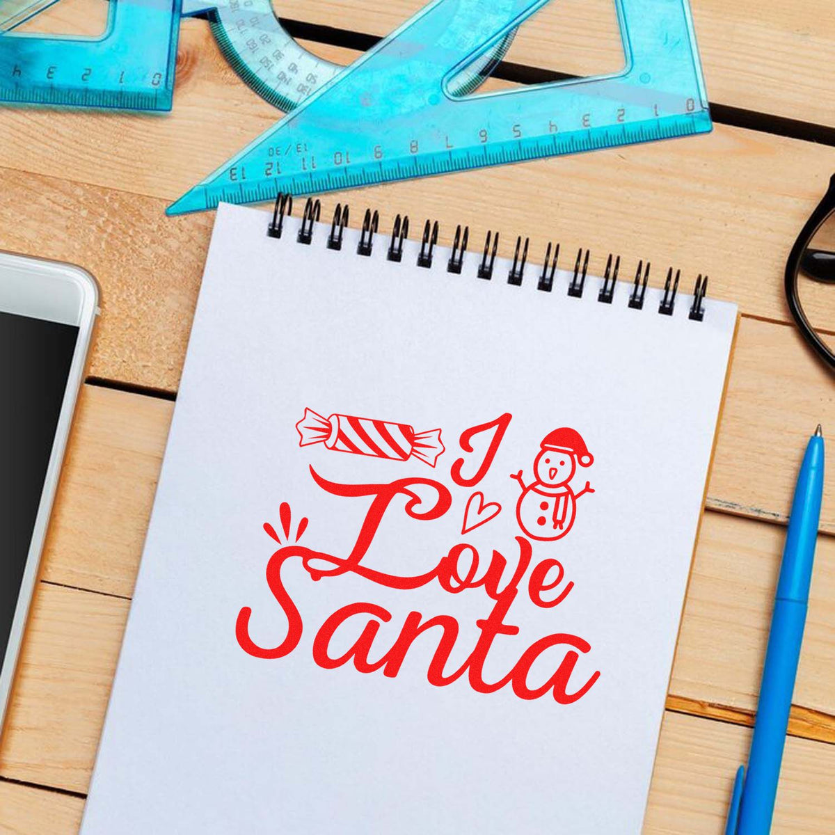 Red Love Santa stamp design with candy cane and snowman on white notepad, beside blue rulers, pen, and wood desk.
