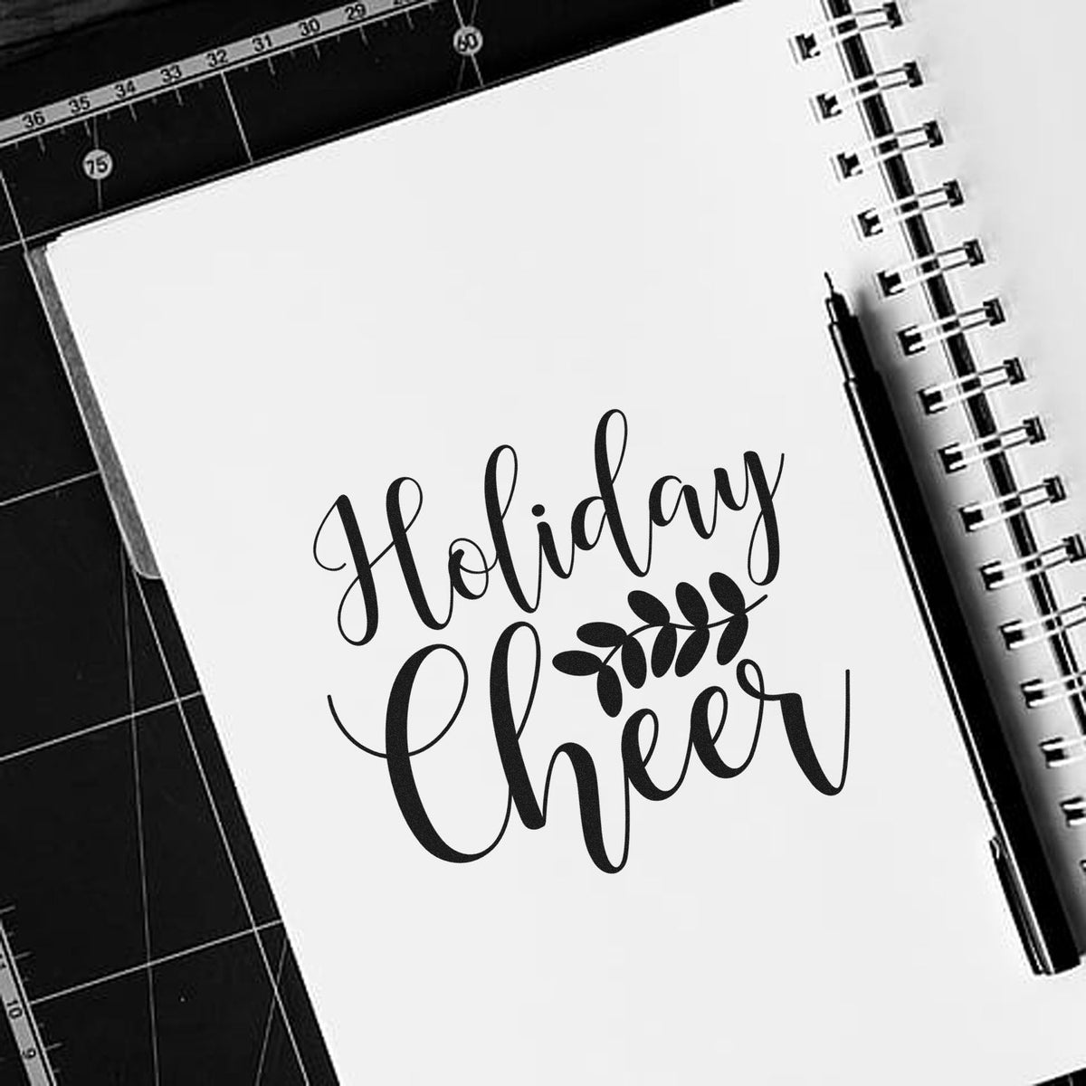 Black holiday cheer stamp and seal on a white spiral notebook, with a black pen on a dark cutting mat.
