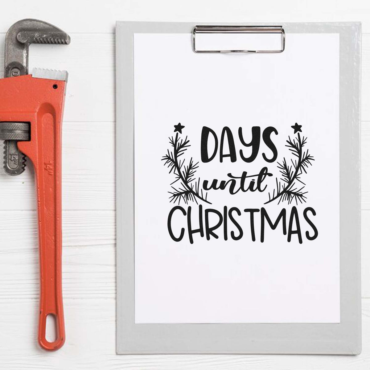 Black Days until Christmas stamp design on clipboard with pine wreath, beside orange pipe wrench.
