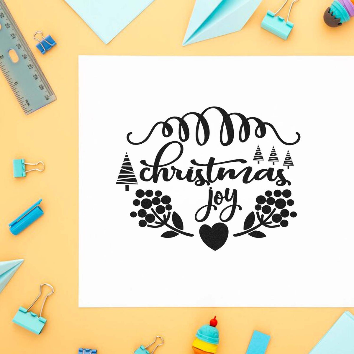 Black Christmas Joy stamp and seal design with trees and heart on white paper, on a yellow desk with pastel blue stationery.