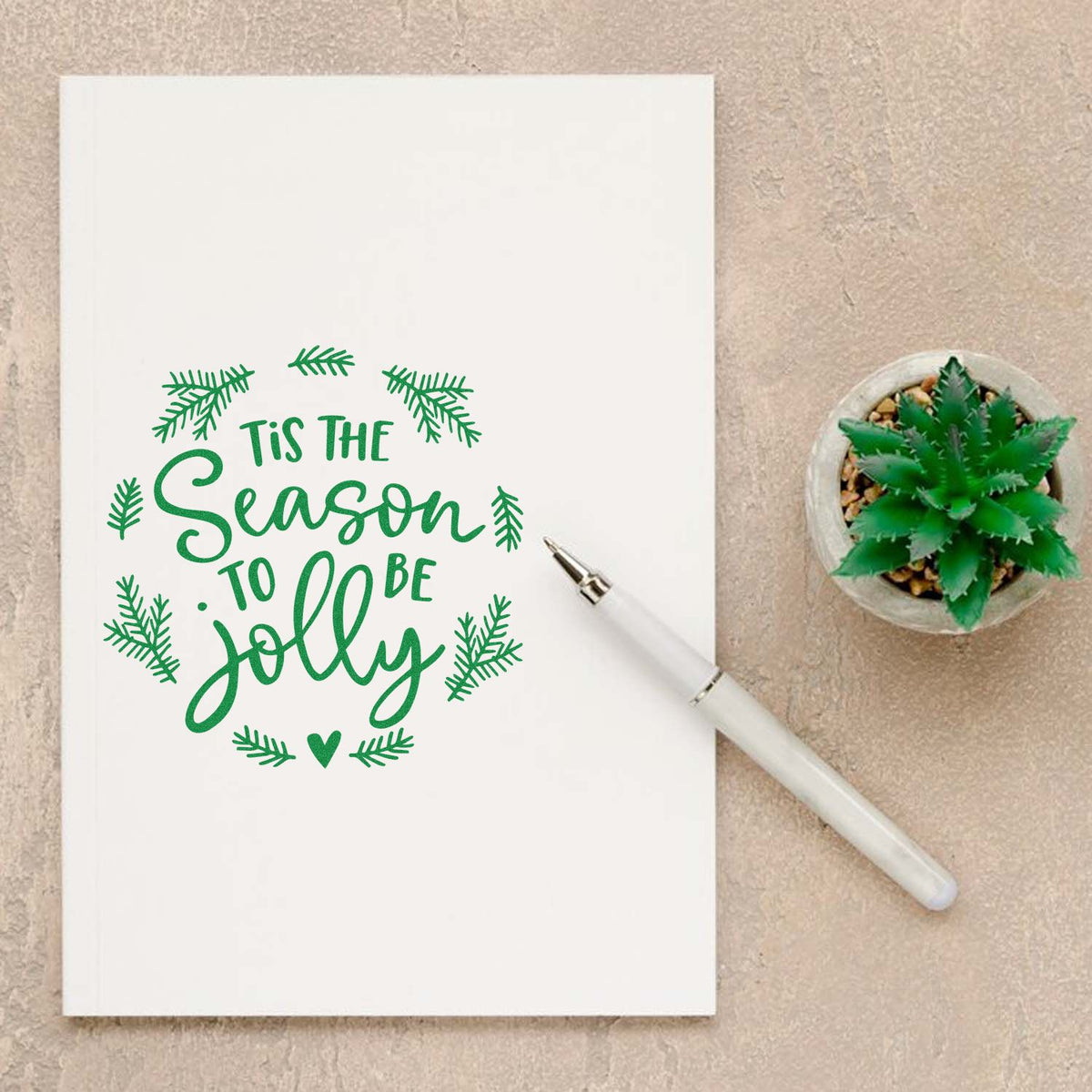 Green holiday stamp and seal reading "Tis the Season to Be Jolly" with pine sprigs, white pen, and succulent.