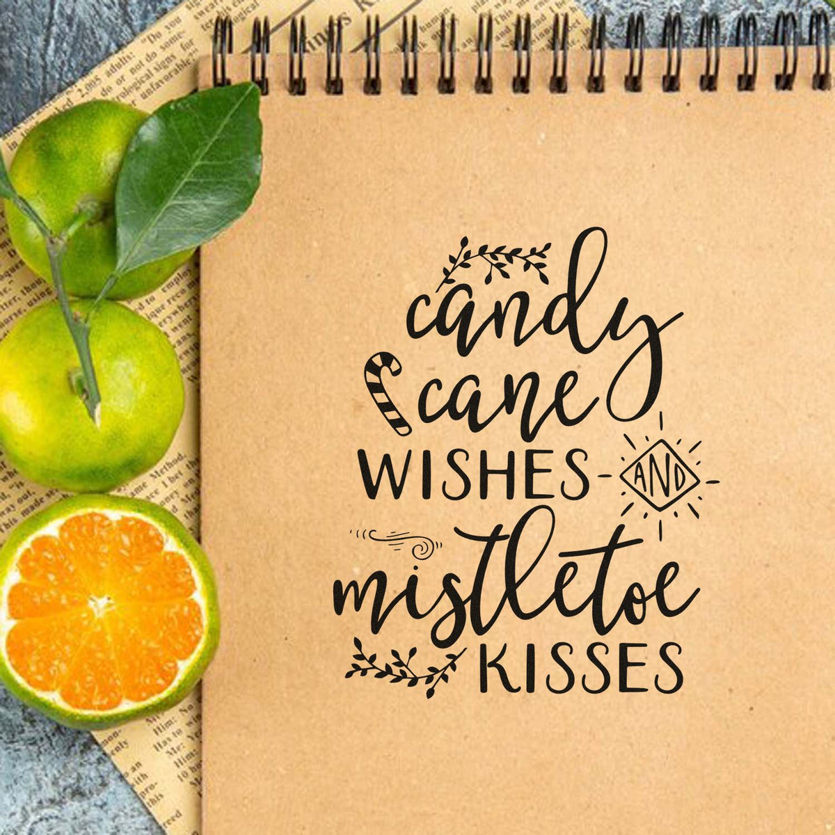 Kraft spiral notebook with black stamp-and-seal holiday design, candy cane wishes and mistletoe kisses, and green citrus slices.