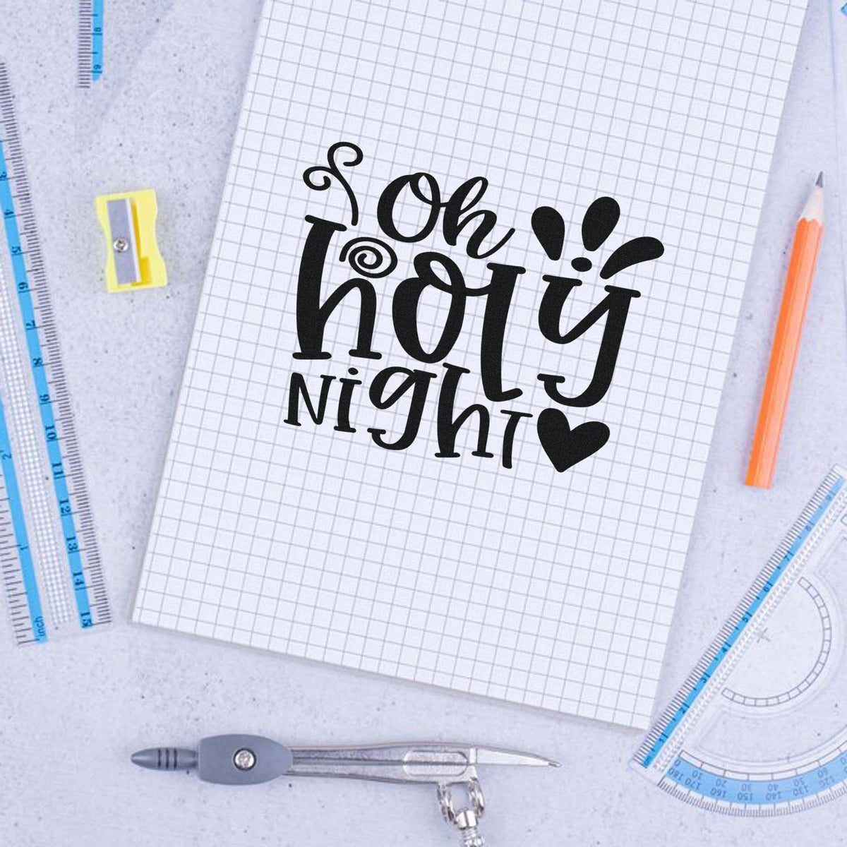 Black Oh holy night stamp and seal design on grid paper, with blue rulers, yellow sharpener, orange pencil, and compass.