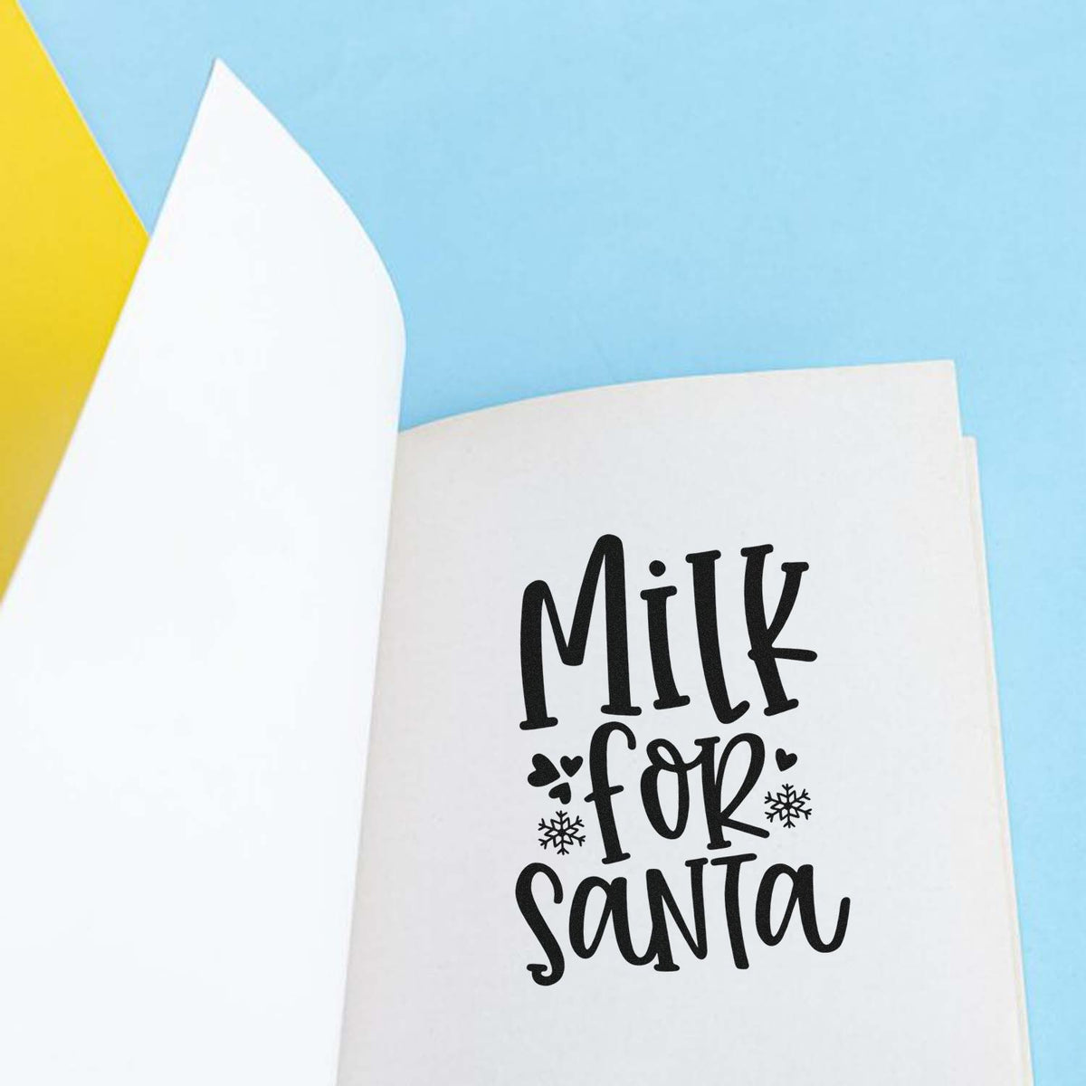 Black Milk for Santa holiday stamp on white paper with snowflakes, against a light blue background and yellow accent.