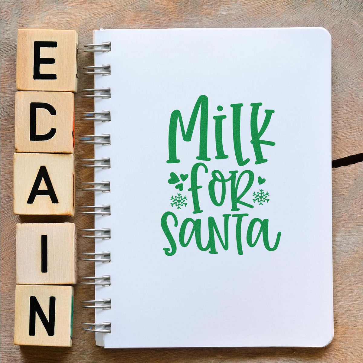 Wooden alphabet stamp blocks next to a spiral notebook with green Milk for Santa seal design and snowflakes.
