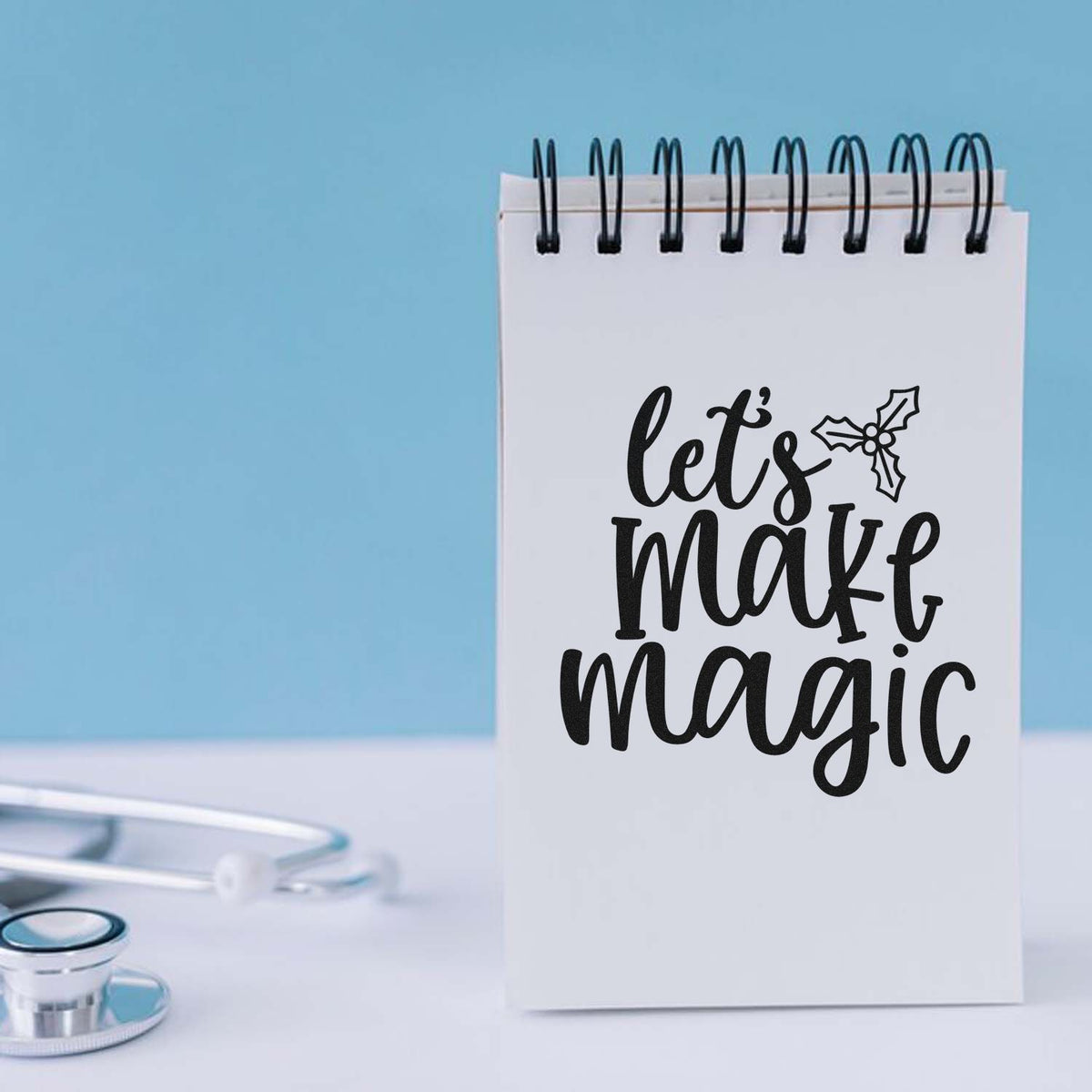 White spiral notepad featuring a black lets make magic stamp and holly seal on a light blue background.