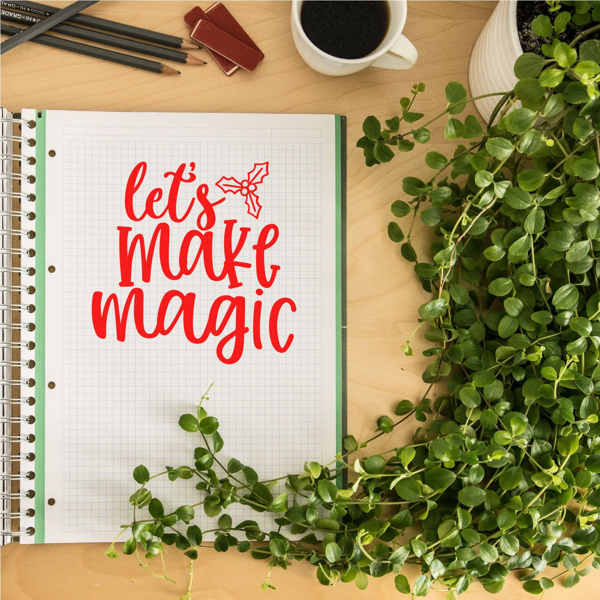 Red holiday stamp reading lets make magic on graph paper notebook, with green plant, coffee cup, and desk tools.
