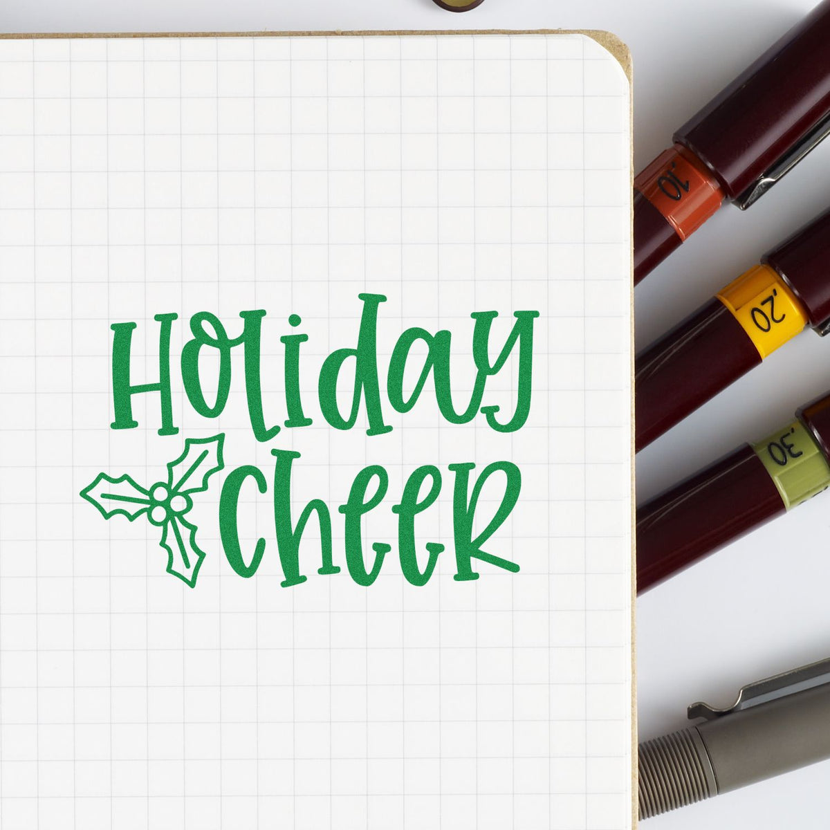 Green Holiday Cheer stamp on a grid notebook with holly, beside maroon stamp and seal pens with colored bands