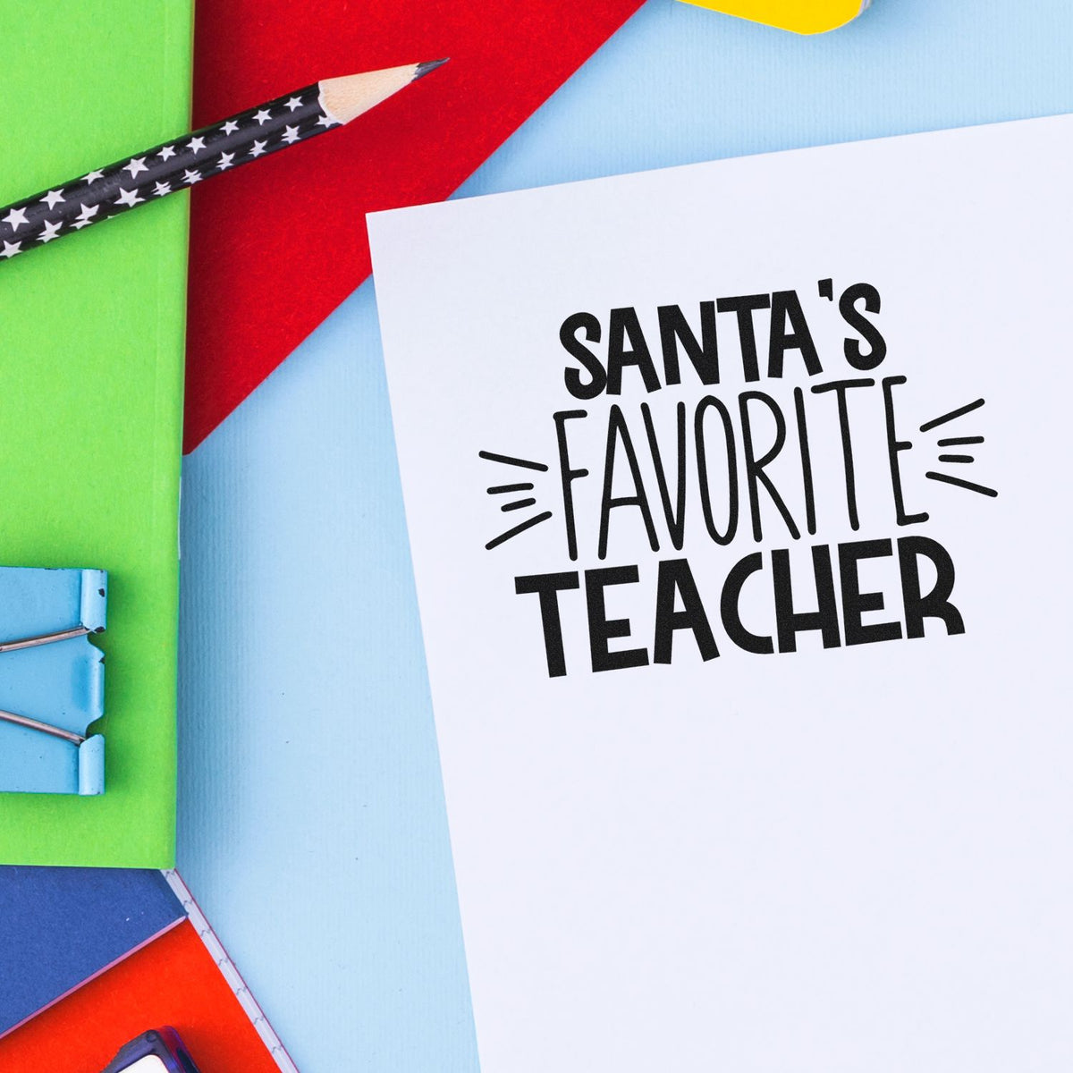 Santas Favorite Teacher stamp on white card with black lettering, surrounded by colorful paper and a star-pattern pencil.