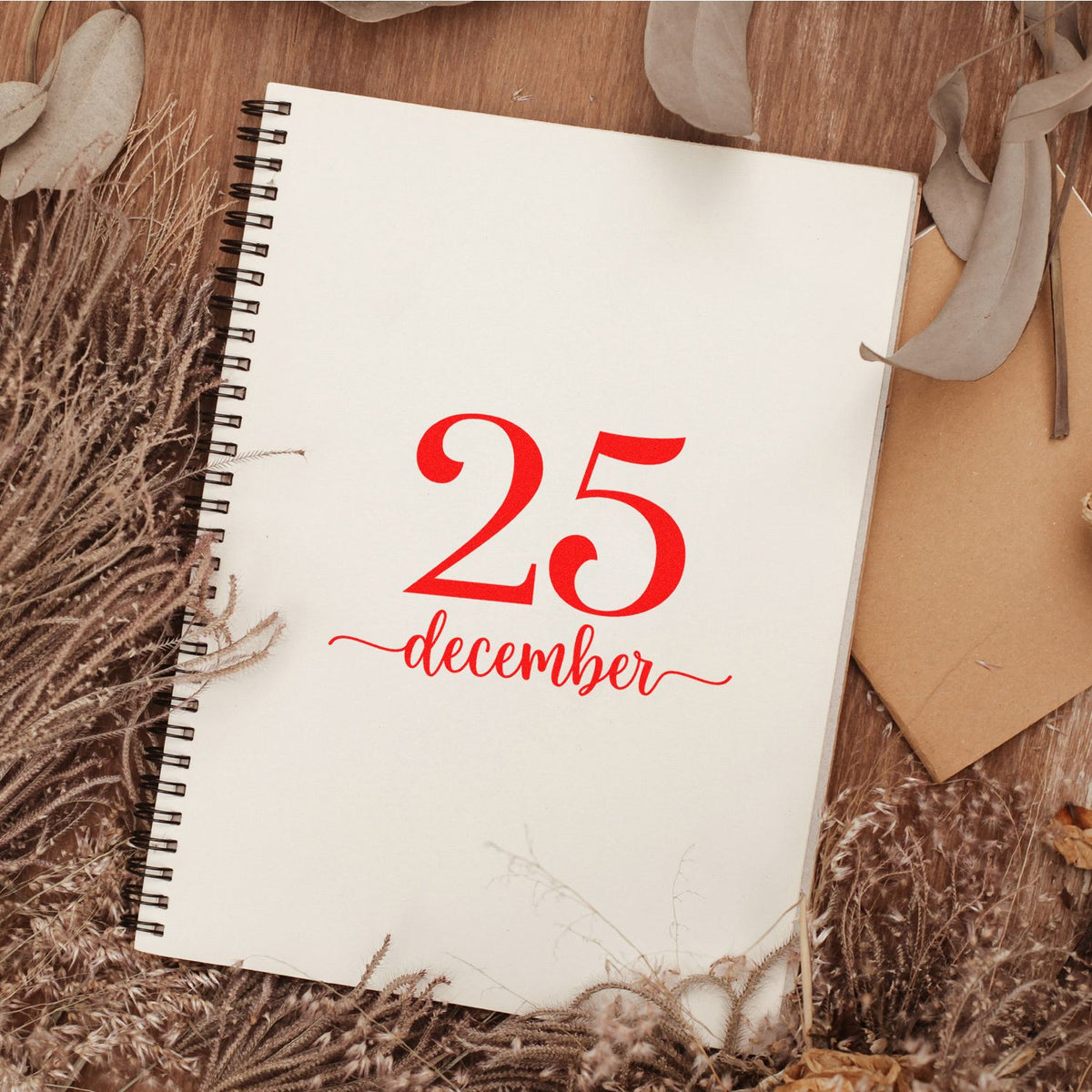 White spiral notebook with a red 25 December stamp and seal design, framed by dried grasses and beige leaves on warm wood.