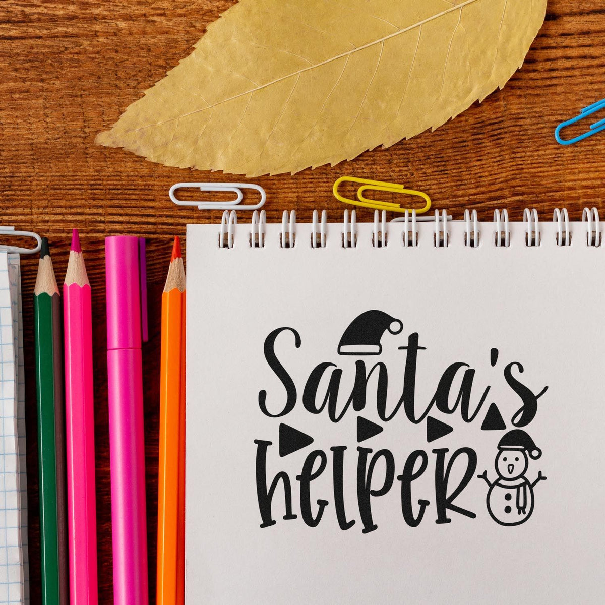 Santas Helper stamp design in black on a white spiral notebook, with colorful pencils, paper clips, and wood tabletop.
