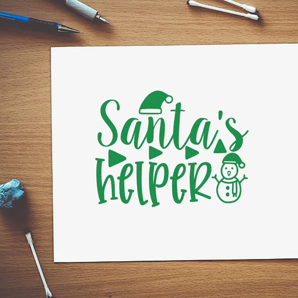 Green Santas helper stamp design on white paper with a Santa hat and snowman, on a wood desk with crafting tools.