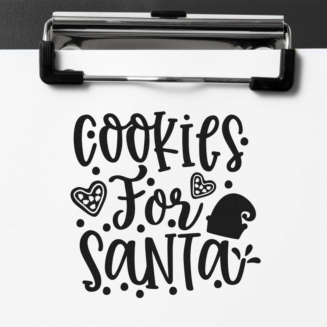 Black stamp and seal design reading Cookies for Santa with hearts and Santa hat on white paper, clipped to a board.