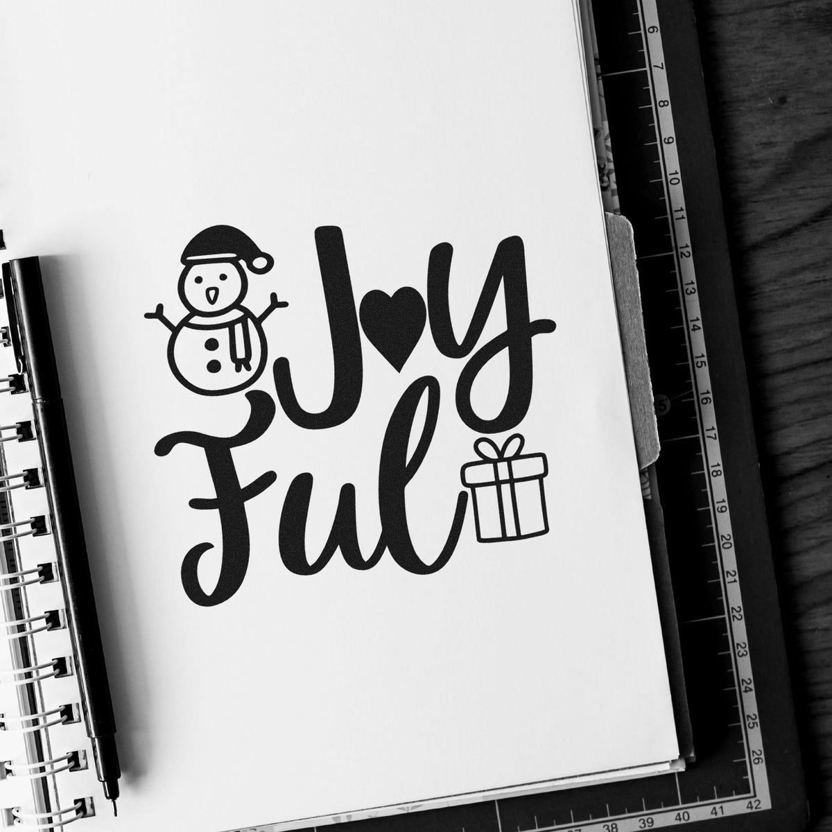Black holiday stamp and seal design reading Joy Ful with snowman and gift on white paper.