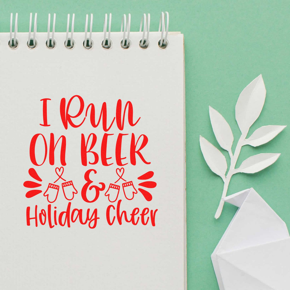 Red holiday cheer stamp and seal design on white spiral notebook, with mint background, white paper leaves, and folded paper accents.