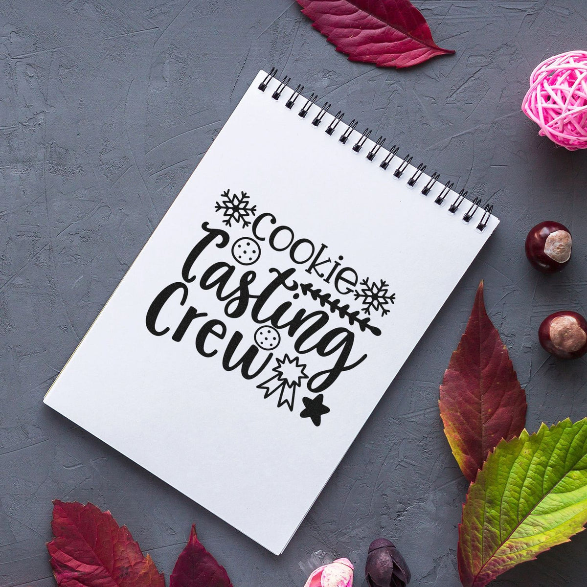 Black cookie-casting crew stamp and seal design on white spiral notebook, with snowflakes, stars, and autumn leaves.