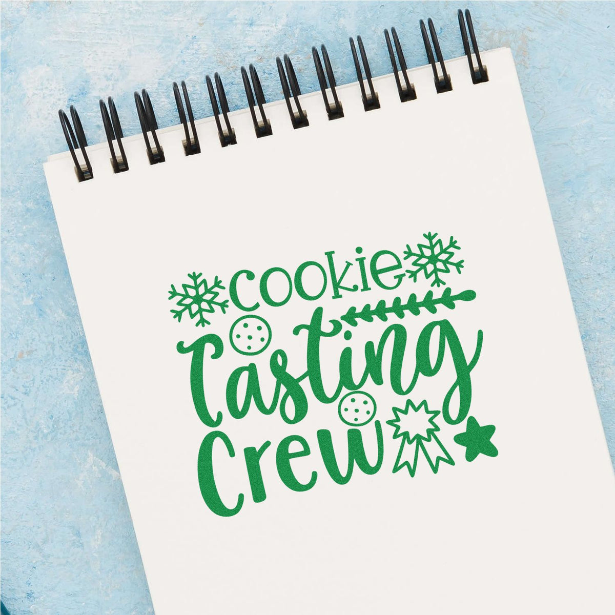 Green cookie tasting crew stamp design on white spiral notepad, with snowflakes, cookie icons, and star accents on blue background.