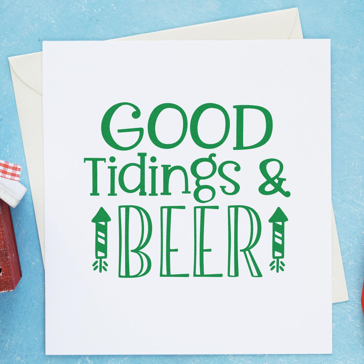Green stamp-style holiday card reading GOOD Tidings & BEER on white paper, with envelopes on a blue background.