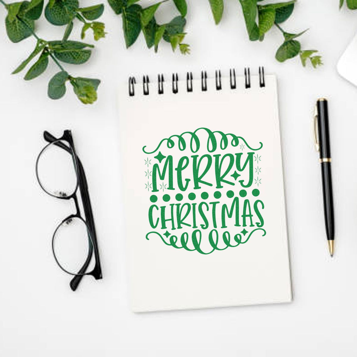 Green Merry Christmas stamp/seal design on white spiral notebook, with swirls, dots, pen, glasses, and greenery.