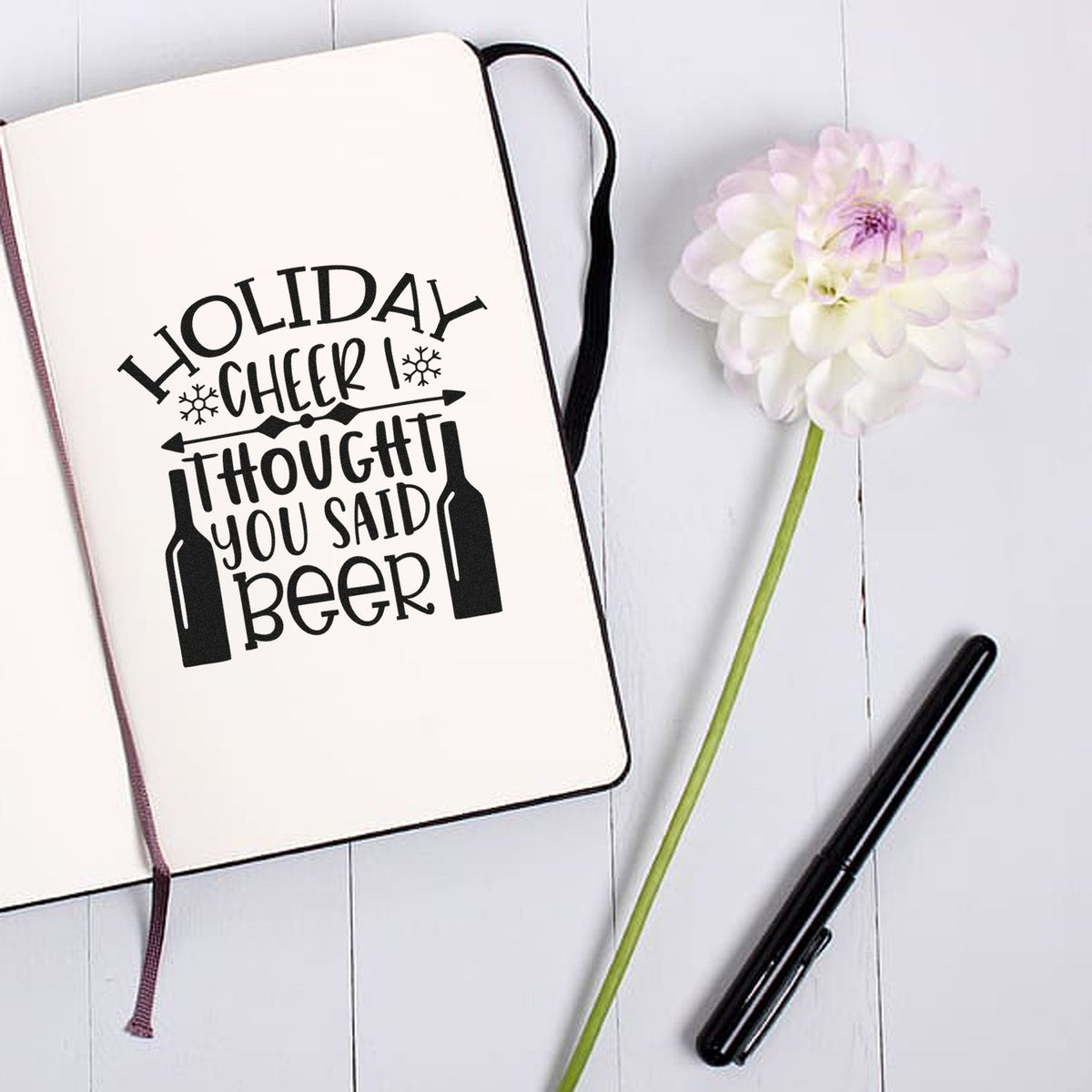 Black holiday beer stamp/seal design on cream notebook, with pink flower, black pen, and white wood background.
