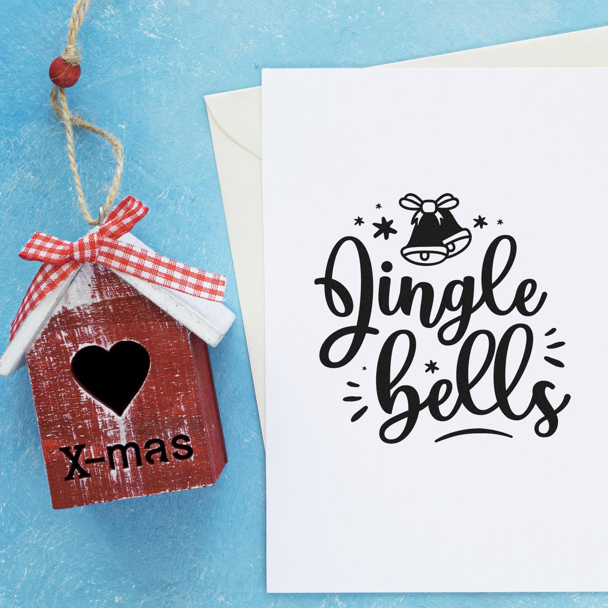 Rustic red wooden birdhouse ornament with heart cutout and X-mas text beside white card with black jingle bells stamp.