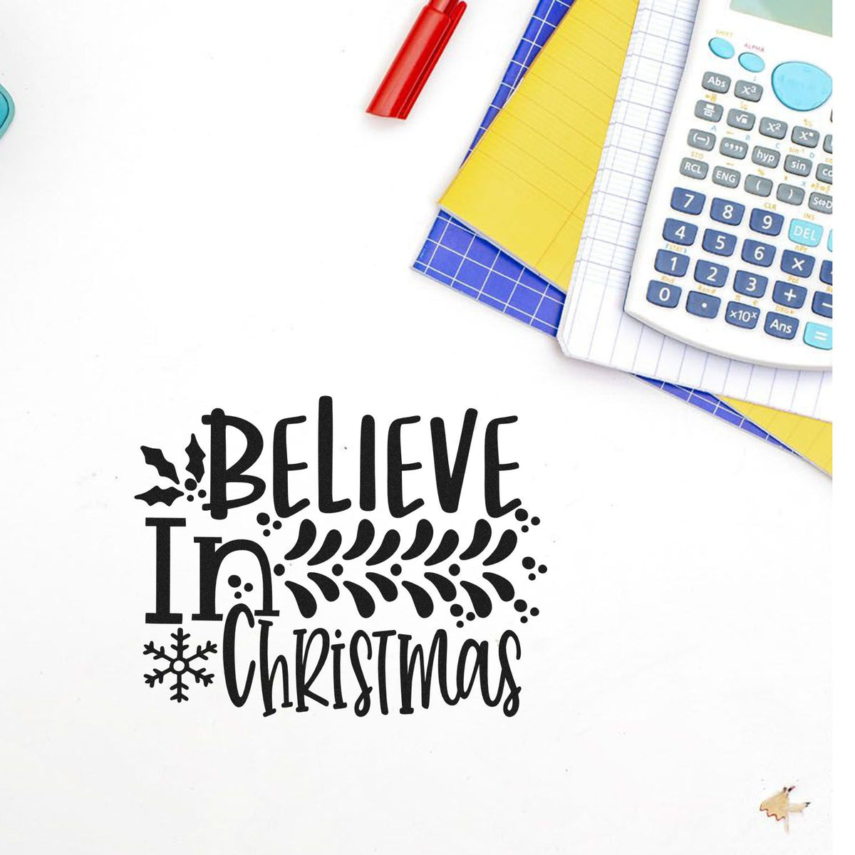 Black Believe In Christmas stamp seal with snowflake, on white background, beside red pen, notebooks and calculator.