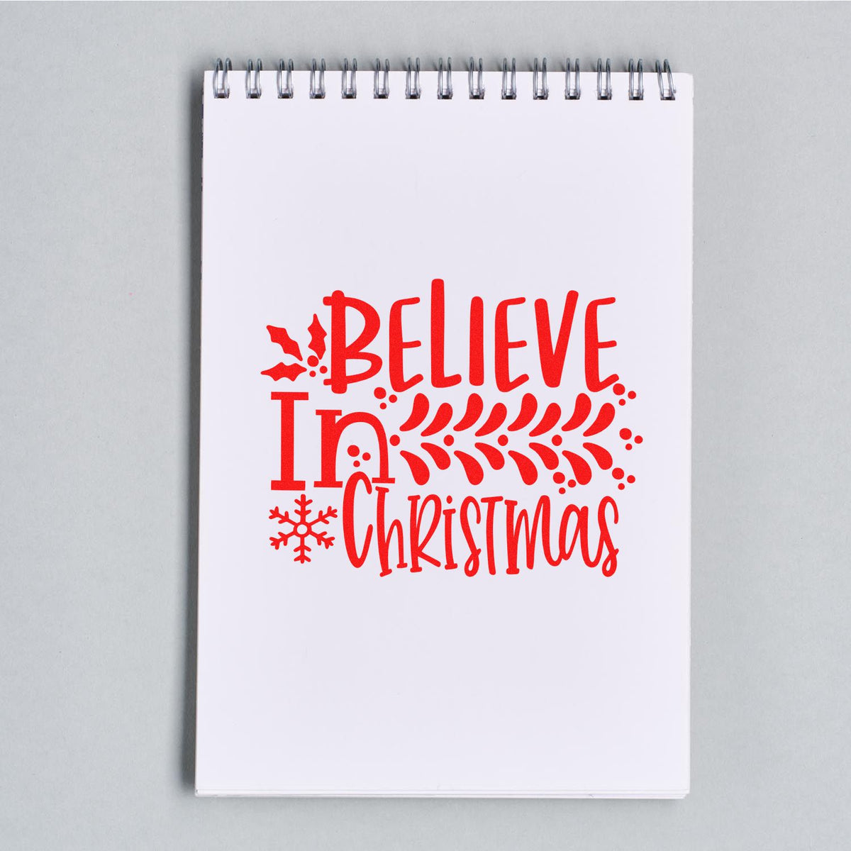 Red holiday stamp and seal design reading Believe in Christmas with holly and snowflake on white spiral notebook.
