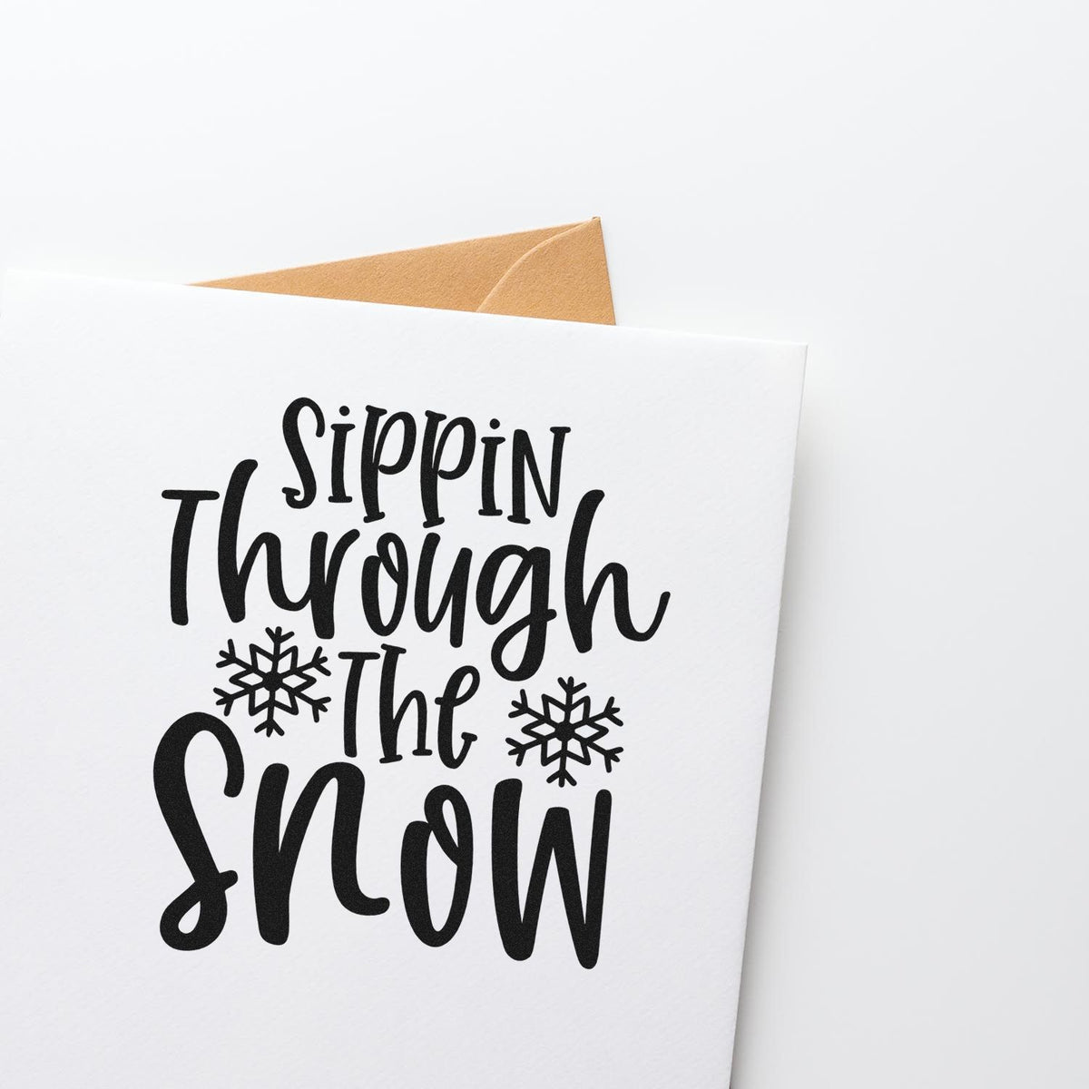 Black holiday stamp reading Sippin Through the Snow with snowflakes on a white card, backed by a brown kraft envelope.