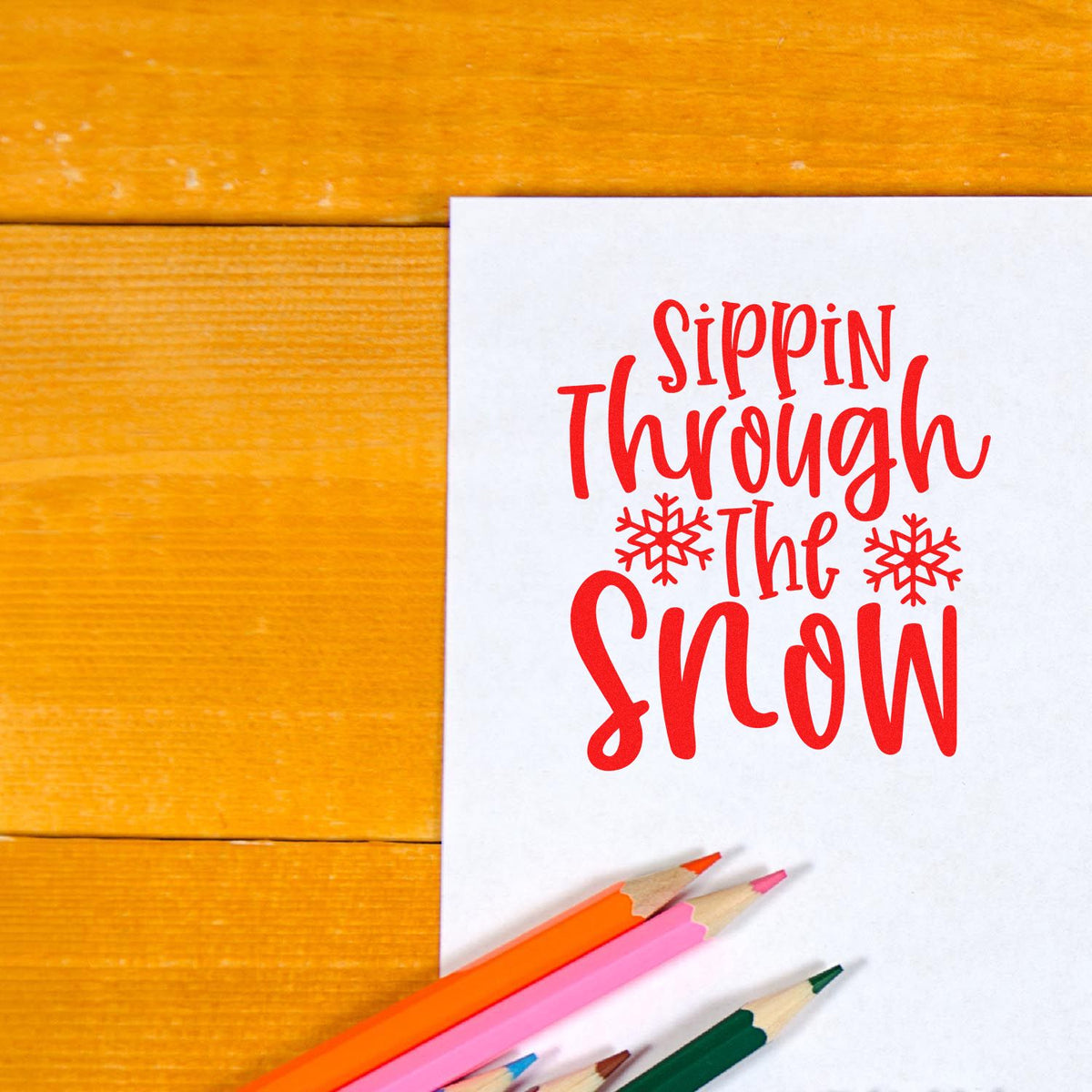 Red holiday stamp/seal reading Sippin Through the Snow with snowflakes on white paper, beside colorful pencils on a wood background.