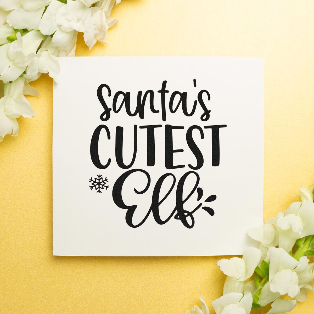 White stamp and seal card with black Santas Cutest Elf lettering, snowflake accent, on soft yellow background with white flowers.