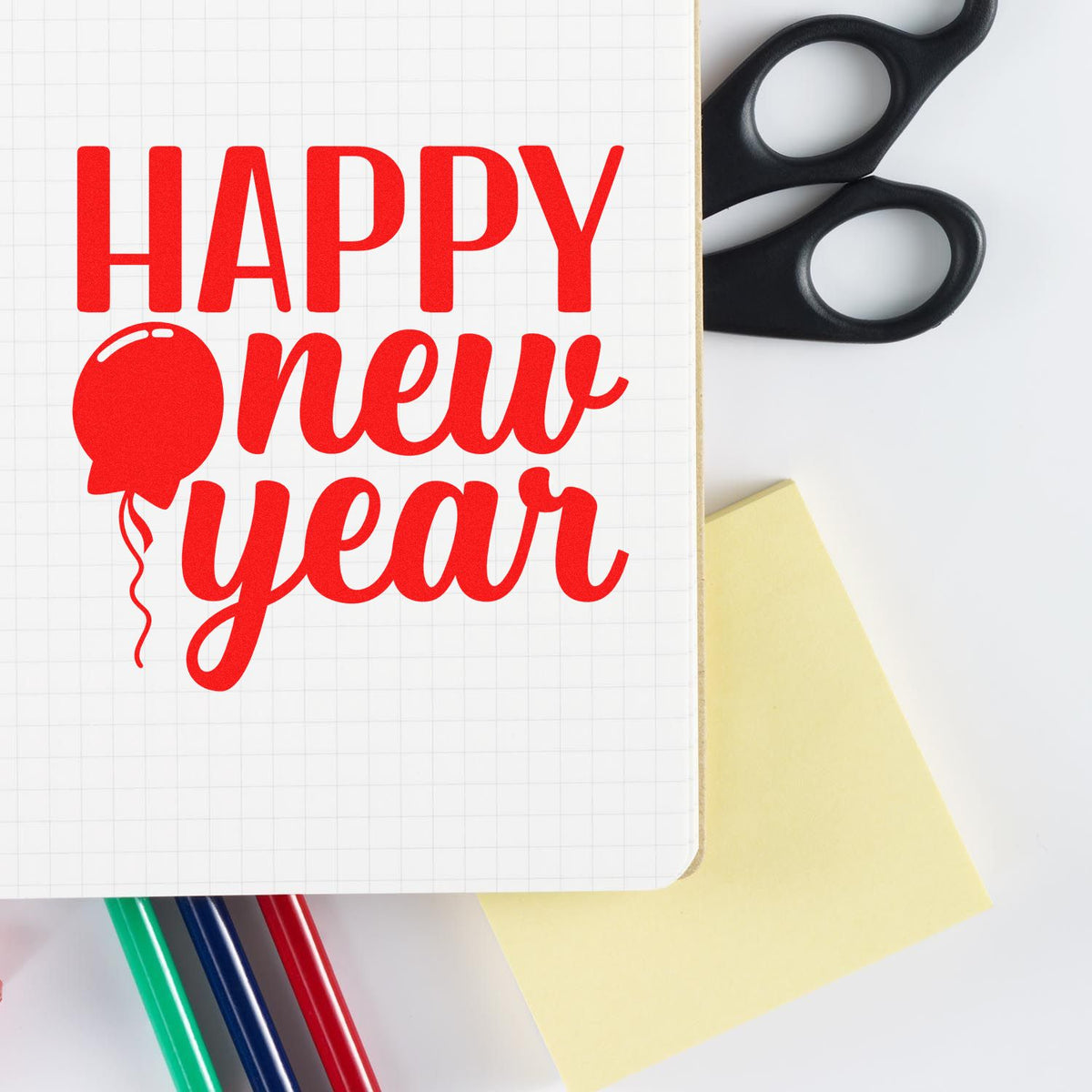 Red Happy New Year stamp design on grid paper with black seal handle, yellow note, and colorful pens.