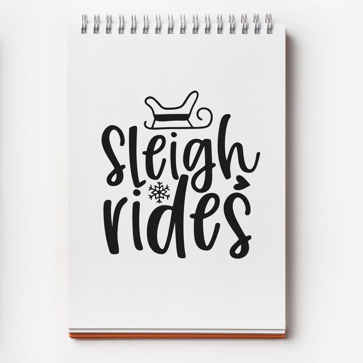 Black holiday stamp design on a white spiral notebook, featuring sleigh and snowflake with Sleigh rides lettering.