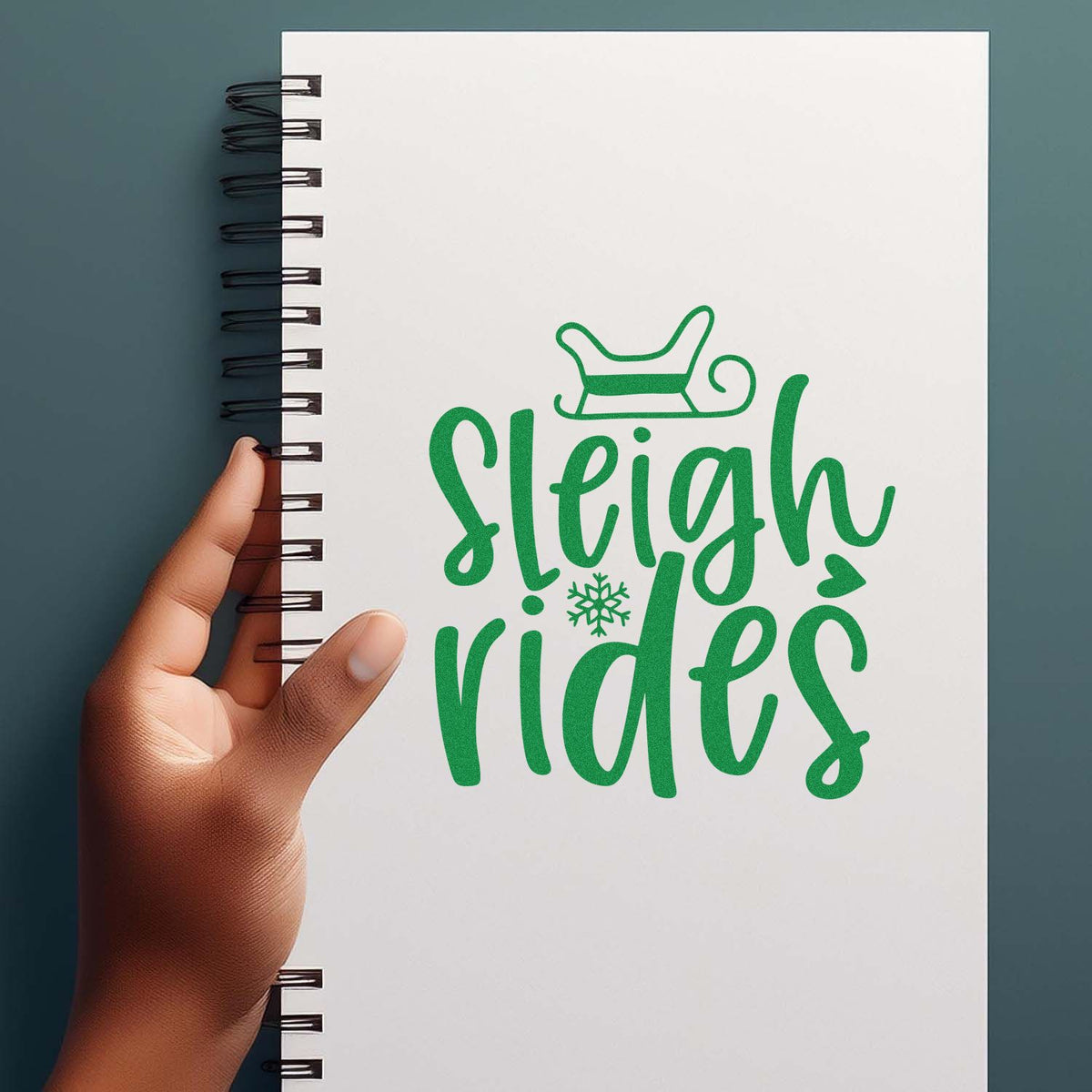 Green Sleigh Rides stamp and seal design on a white spiral notebook, with sleigh and snowflake, held by hand.