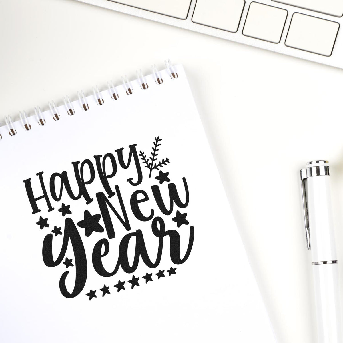 Black Happy New Year stamp on white spiral notebook with stars, beside a white stamp-and-seal pen on a white desk.