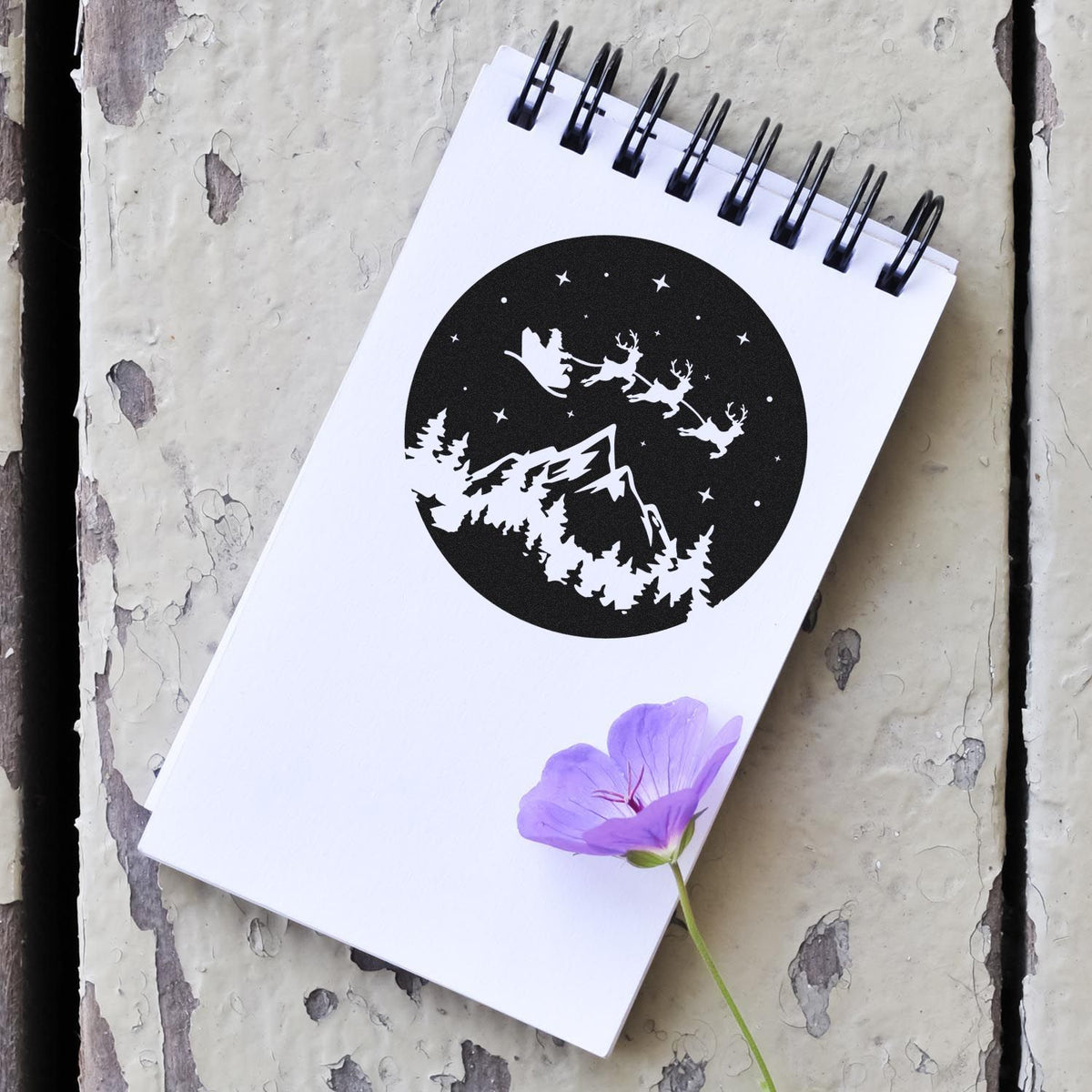 White spiral notebook featuring a black winter stamp seal with Santa sleigh, reindeer, mountains, pine trees, and a purple flower.