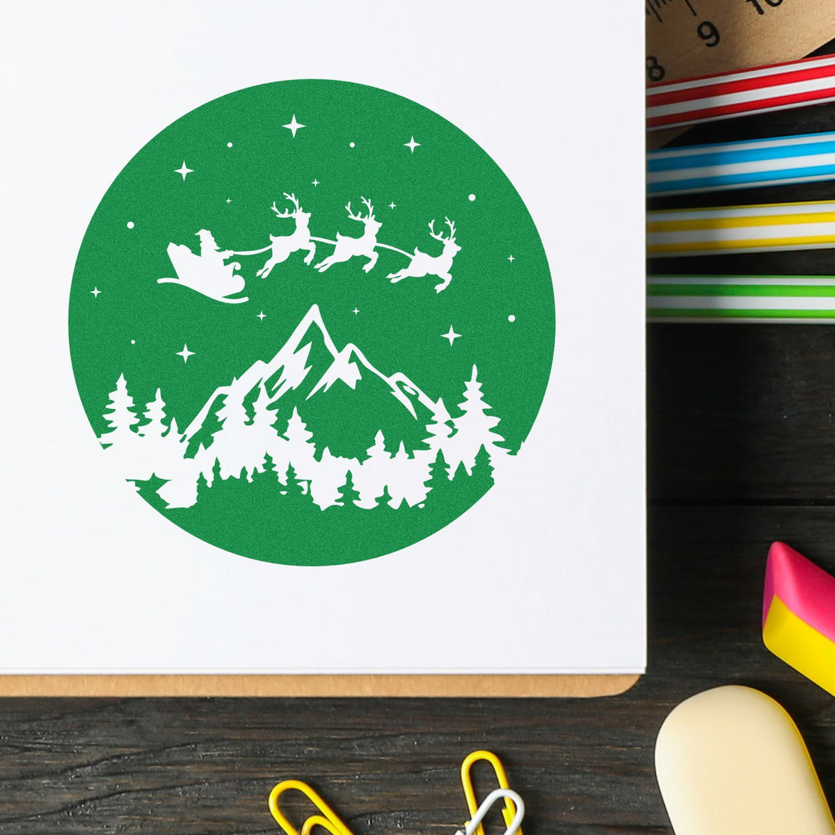 Green circular stamp seal with white Santa sleigh, reindeer, mountain, pine trees and stars on white paper
