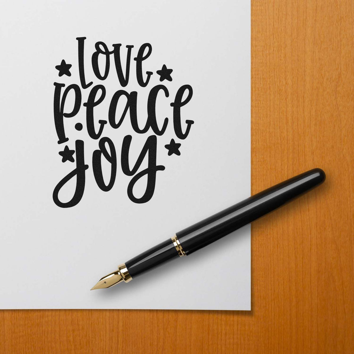 Black custom rubber stamp design reading love peace joy on white paper, with black fountain pen on a wooden desk.