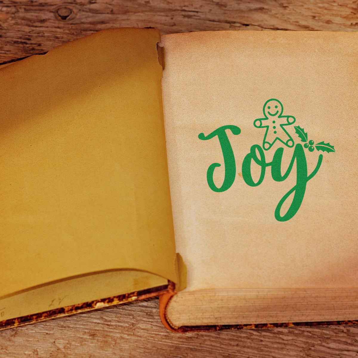 Open vintage book with green Joy stamp and gingerbread seal on cream page, set against rustic wood.