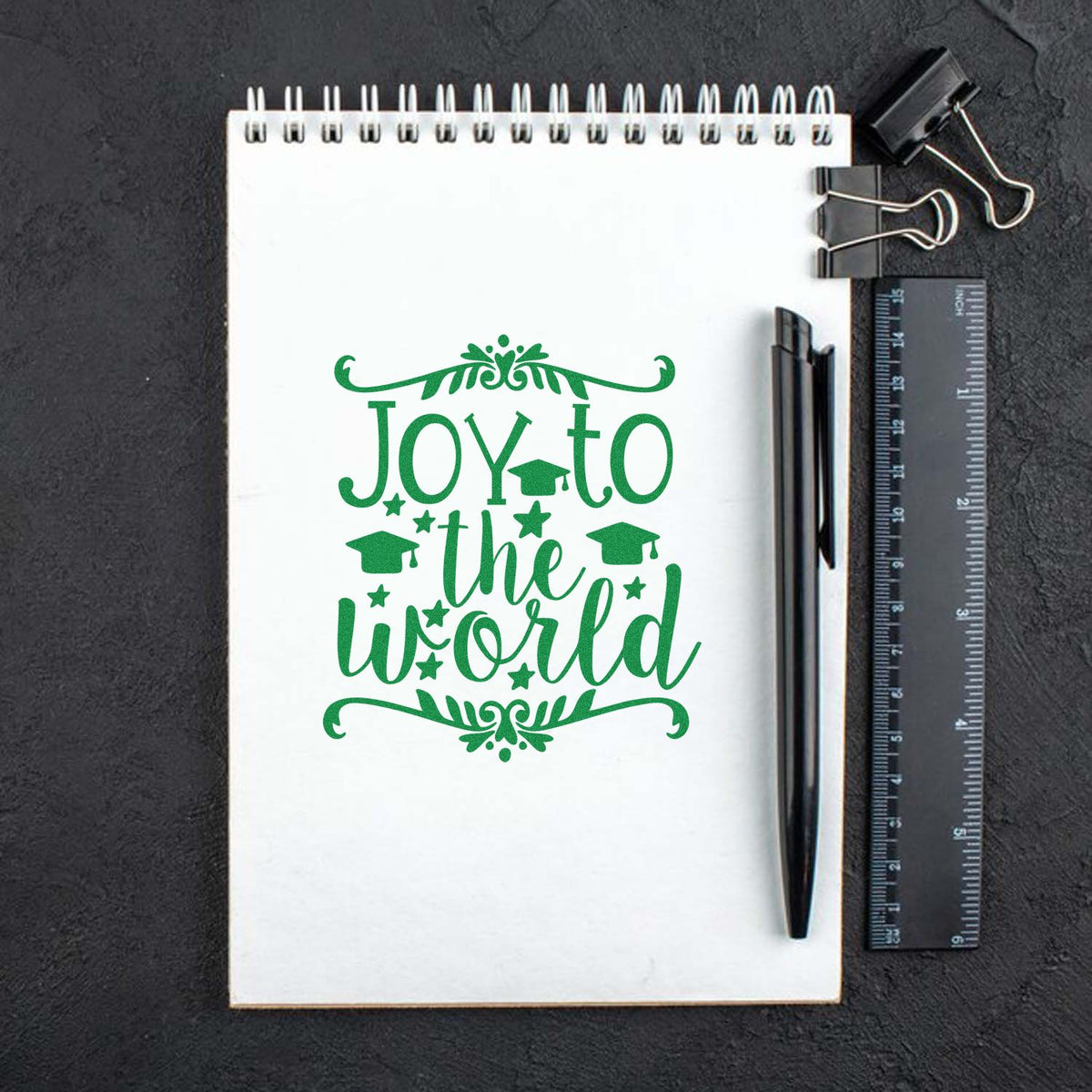 Green graduation stamp reading Joy to the World on a white spiral notebook, with black pen, ruler, and binder clips on a dark background.