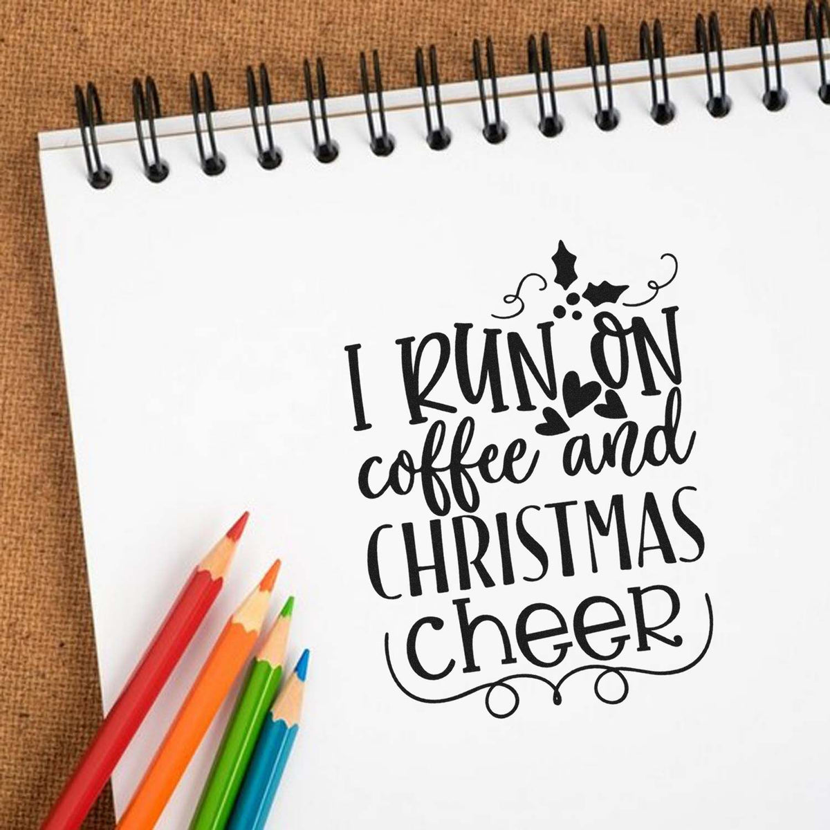 Decorative black holiday stamp and seal on white notebook reading I run on coffee and Christmas cheer, with colorful pencils.