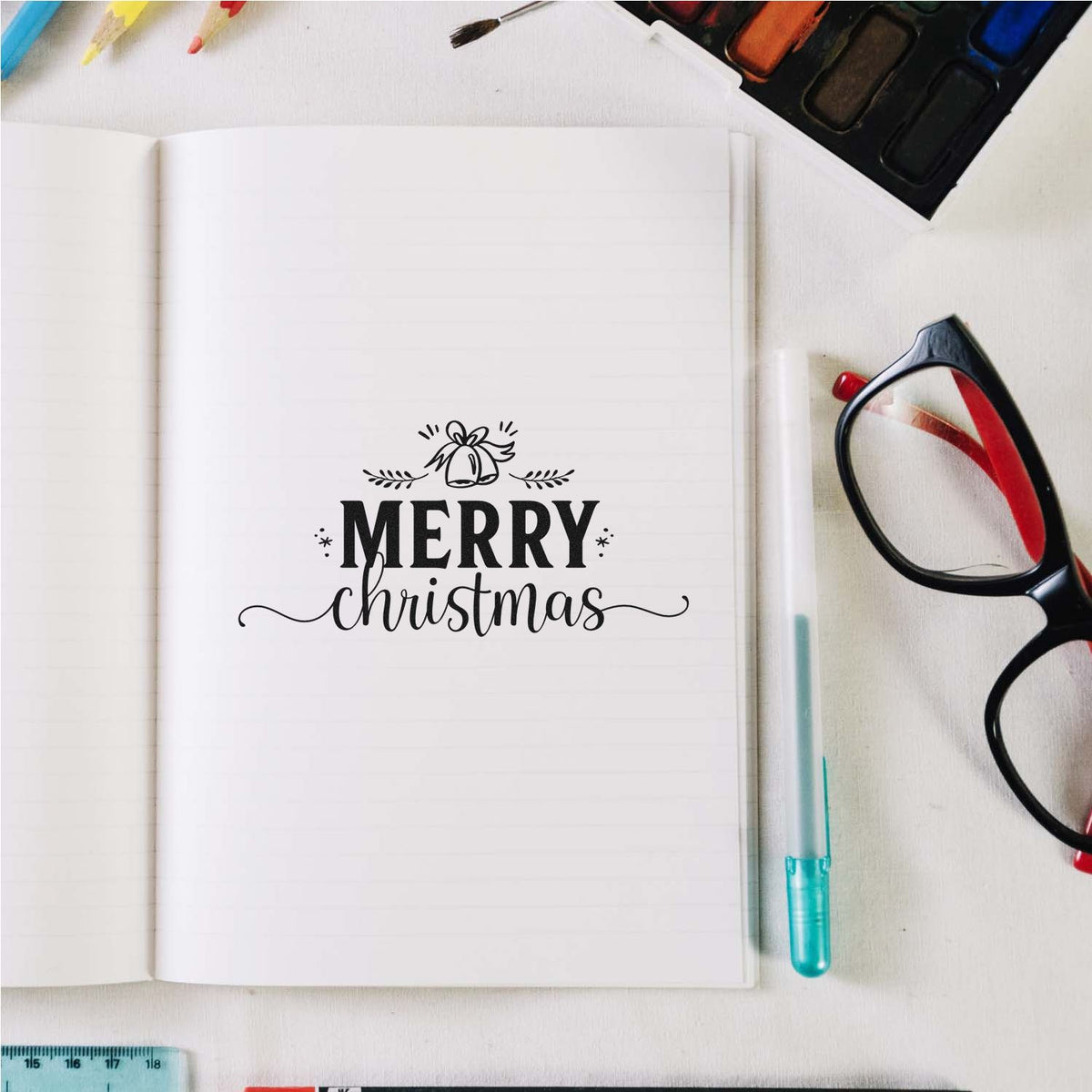Merry Christmas stamp and seal in black on a white notebook page, with craft supplies, glasses, and watercolor palette.