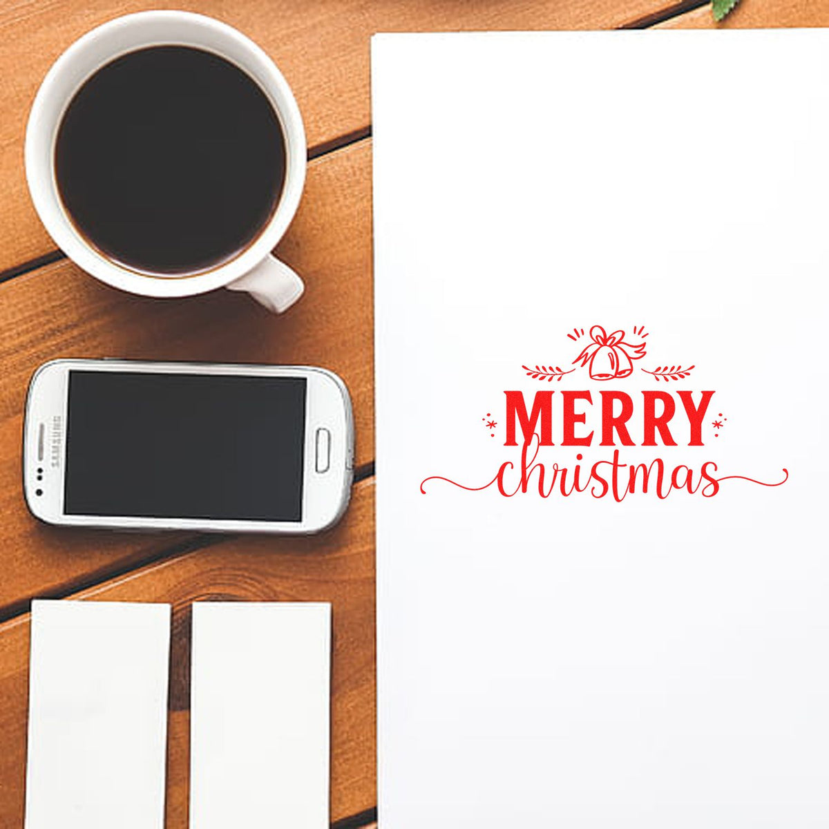 Red Merry Christmas stamp on white paper, with seal tools, coffee cup, and smartphone on a wooden desk.