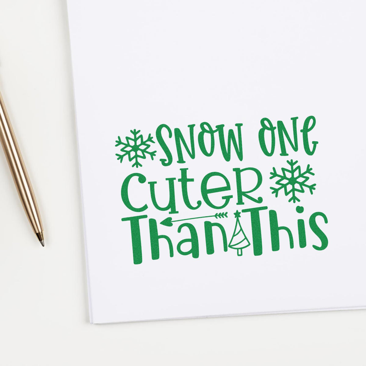 Green holiday stamp reading Snow One Cuter Than This with snowflakes on white paper, beside a gold pen.