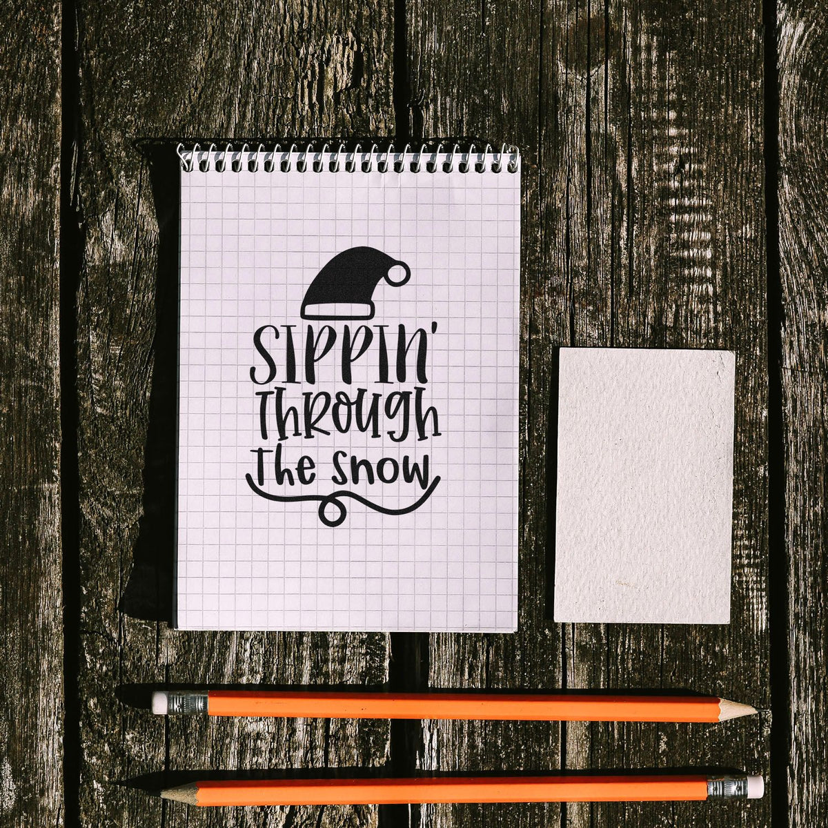 Black holiday stamp and seal design on white grid notepad, with blank card and orange pencils on weathered wood.